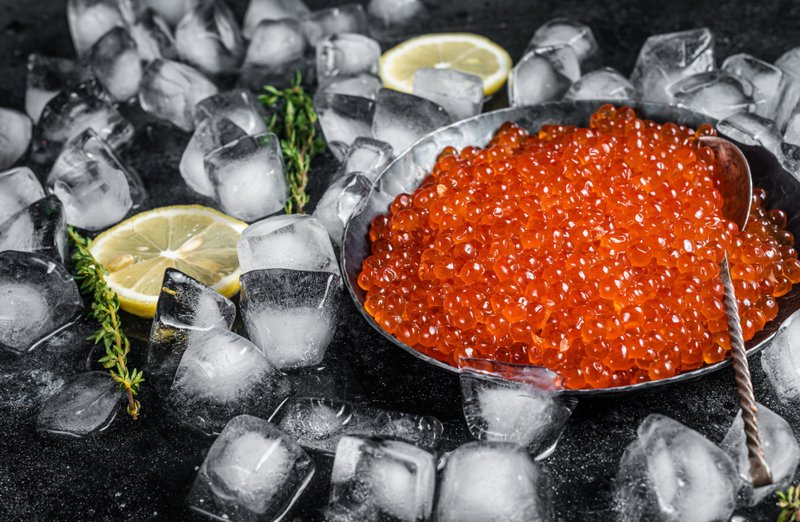 Bowl of bright red salmon caviar surrounded by ice cubes, lemon slices, and thyme sprigs.